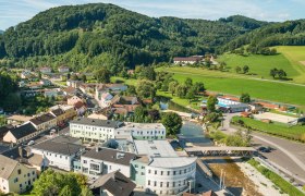 Aerial view of a small village with river, bridge and surrounding hills.