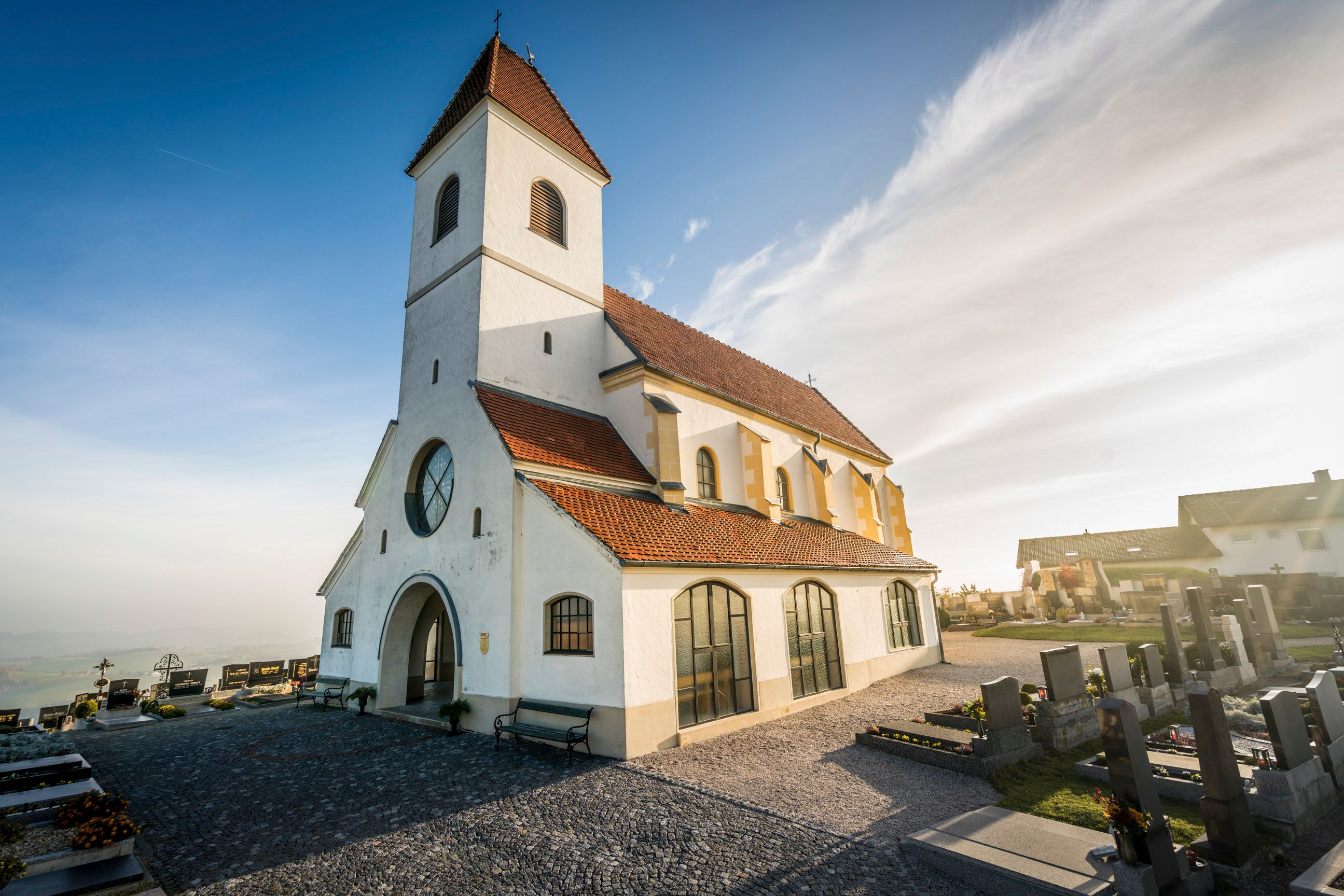 A white church with a red tiled roof and a cemetery in the foreground in sunny weather.