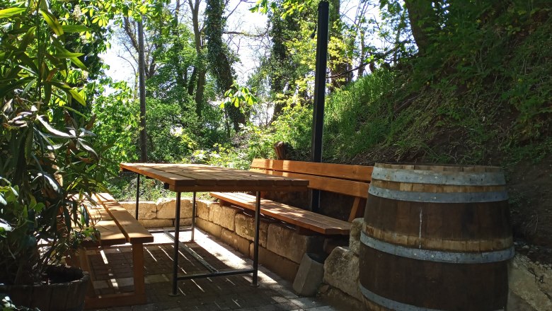 A shaded outdoor seating area with wooden benches and a table, surrounded by trees and plants.