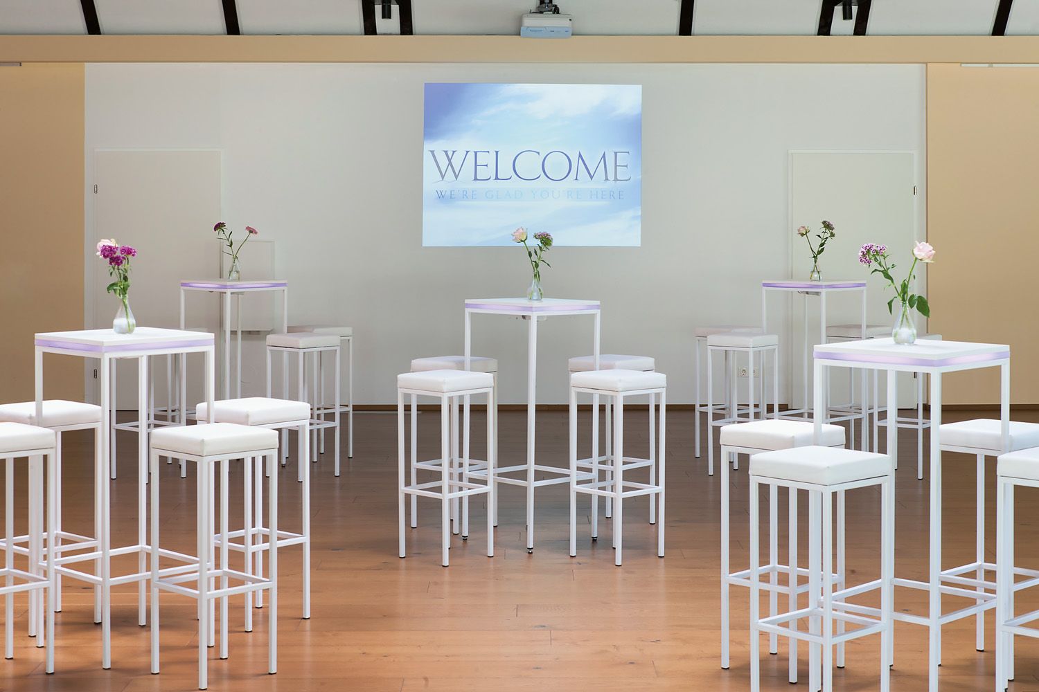 Seminar room with white bar tables and stools, vases of flowers and a screen with the inscription 'Welcome'.