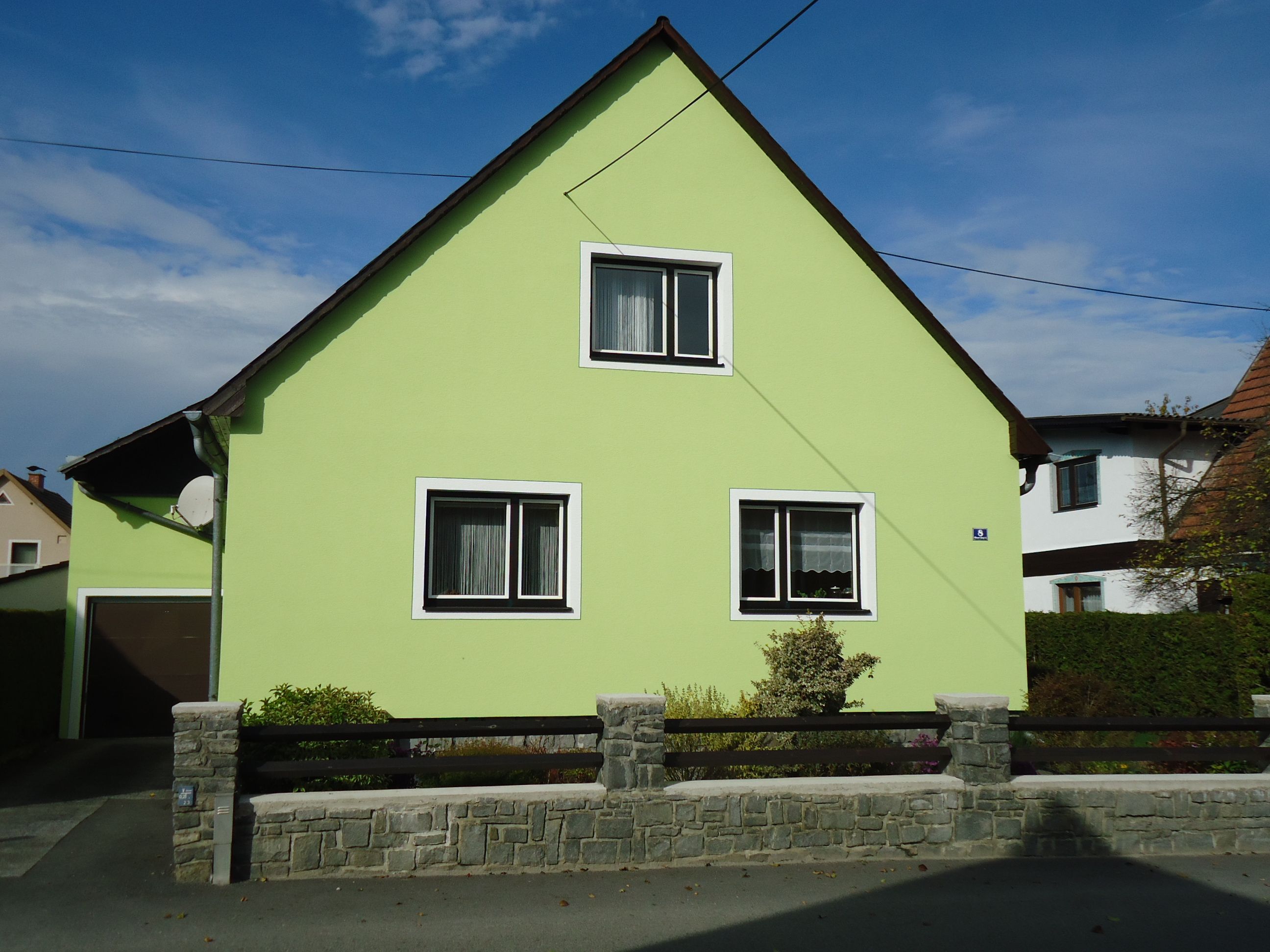 A light green house with a pitched roof and two windows, surrounded by a low stone fence.