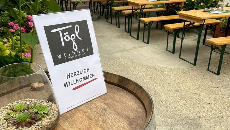 A sign with the inscription 'Weingut Tögl' and 'Herzlich Willkommen' is placed on a wine barrel. Beer tables and plants can be seen in the background.
