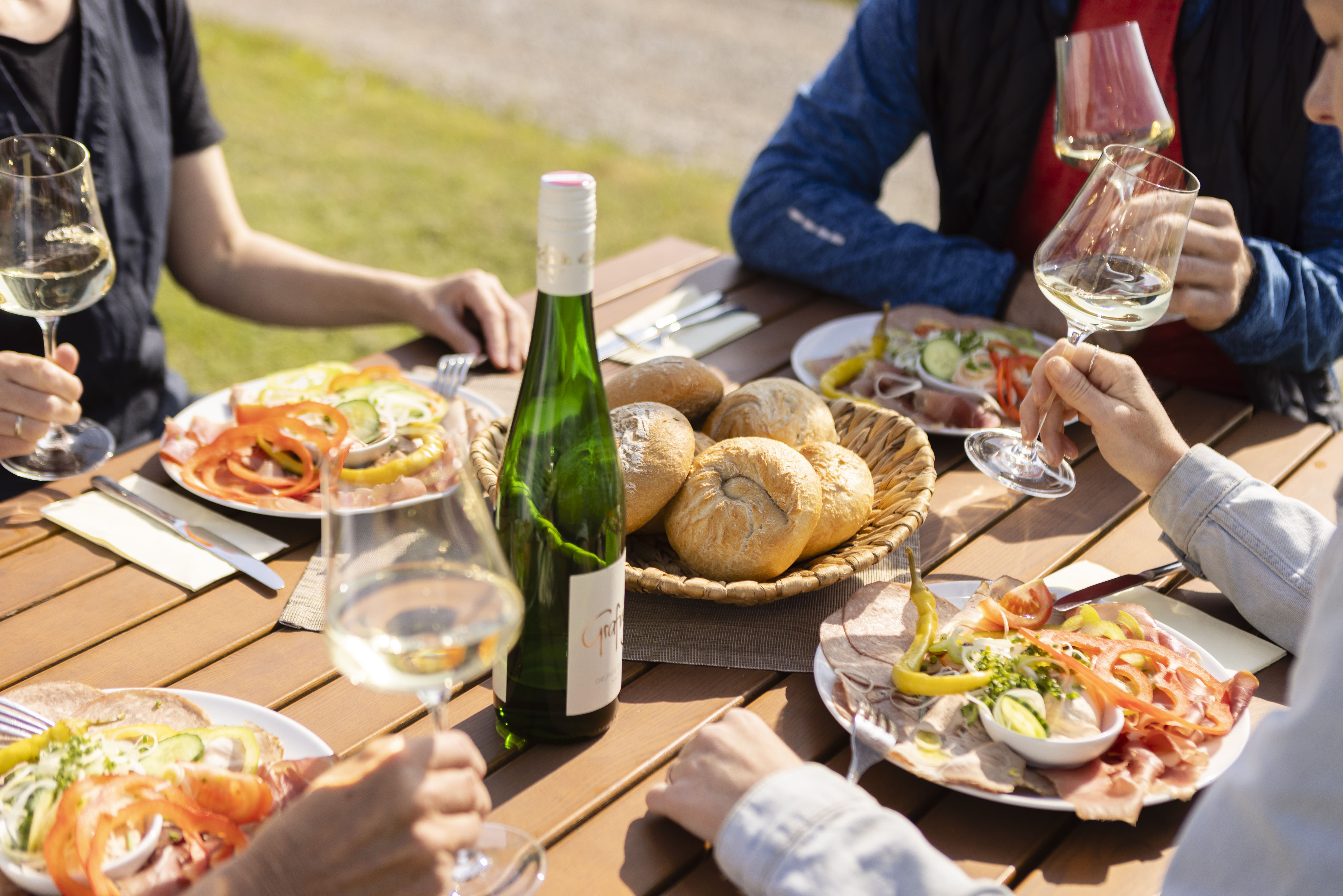 People enjoy wine and food outdoors at a wooden table.