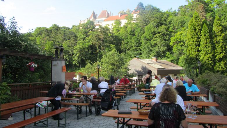 People sit at wooden tables outside with a view of a castle in the background.