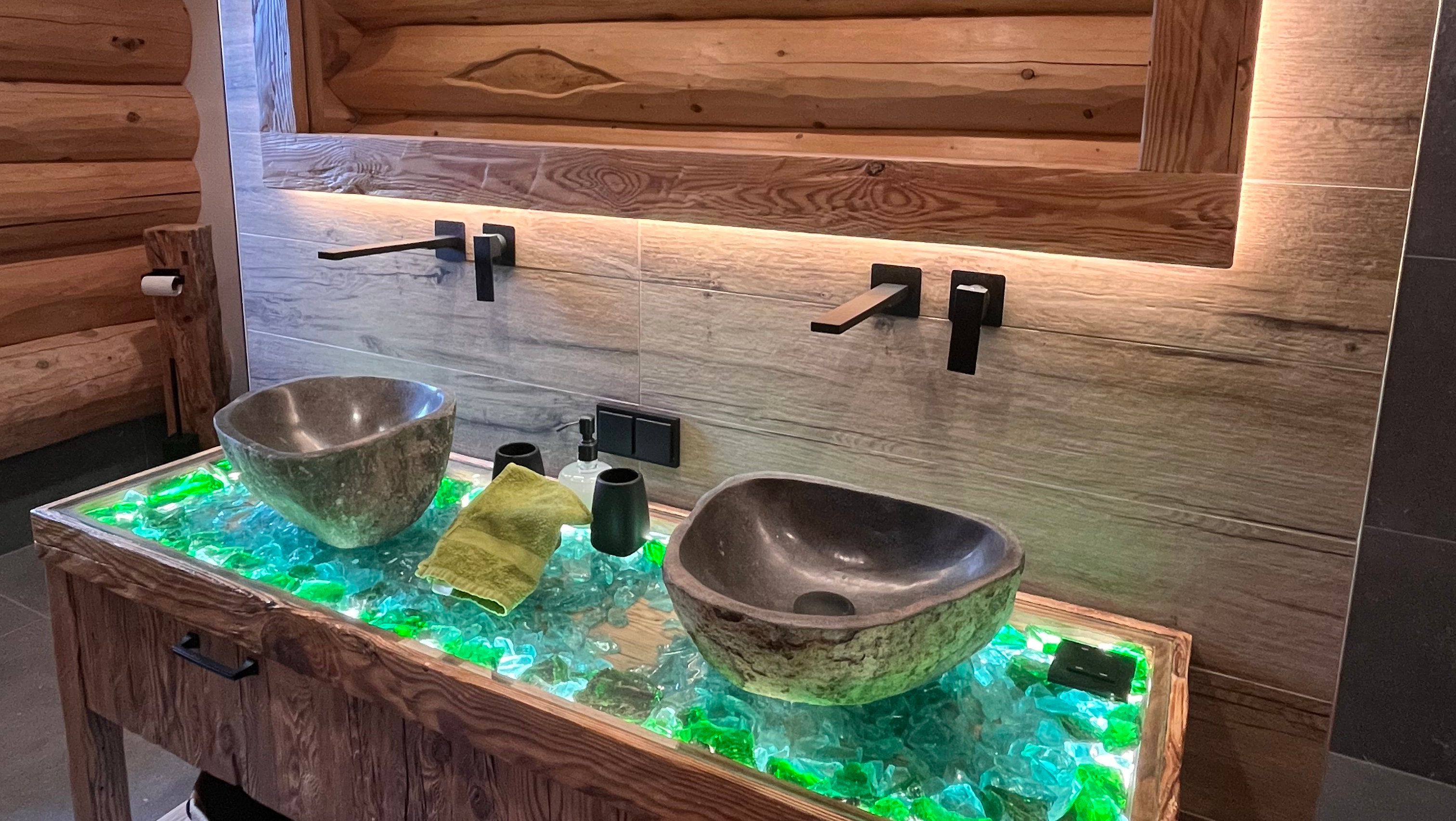 Modern bathroom with two stone washbasins on an illuminated glass table.