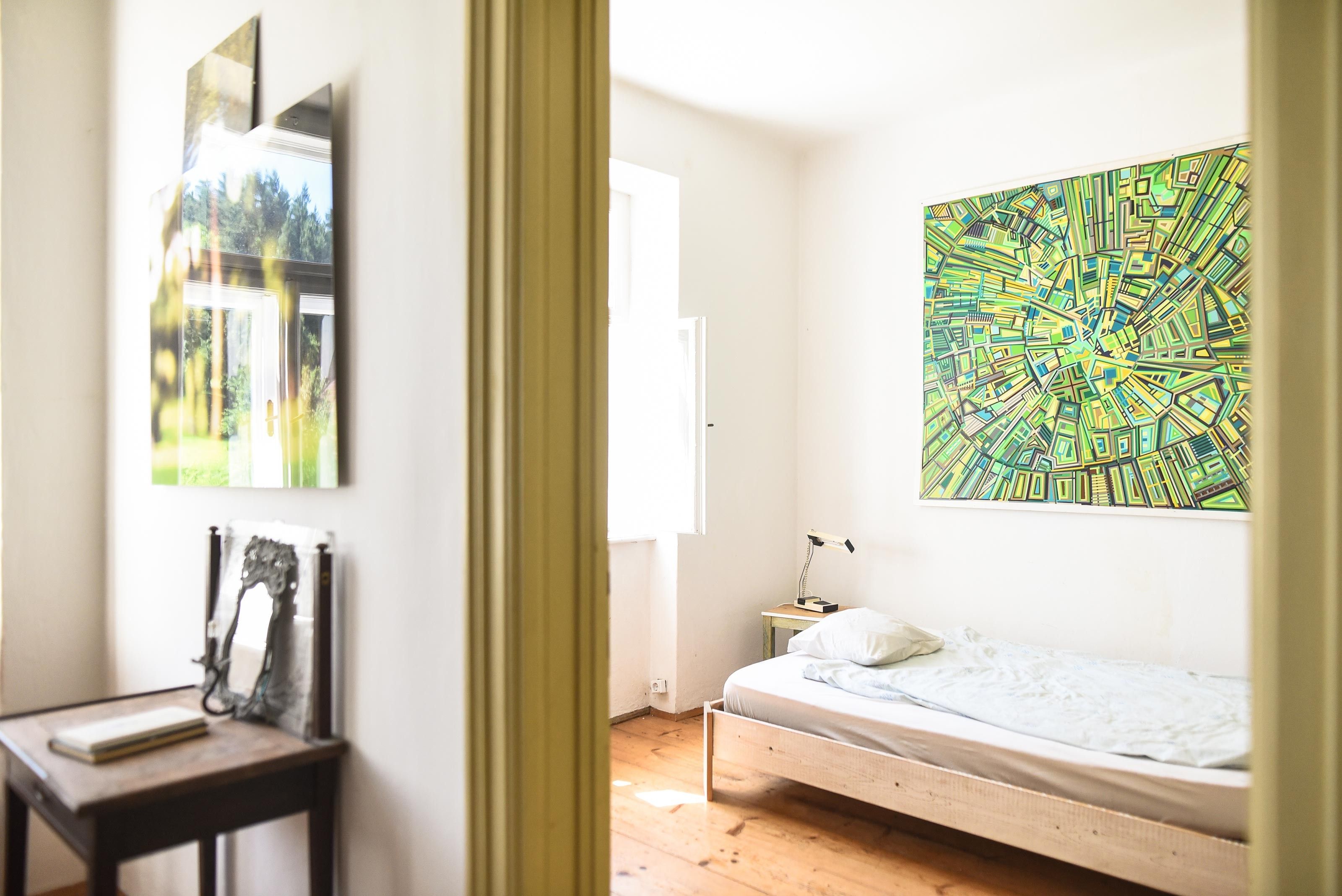 A simply furnished bedroom with a single bed, a small table, and a large, colorful painting on the wall.