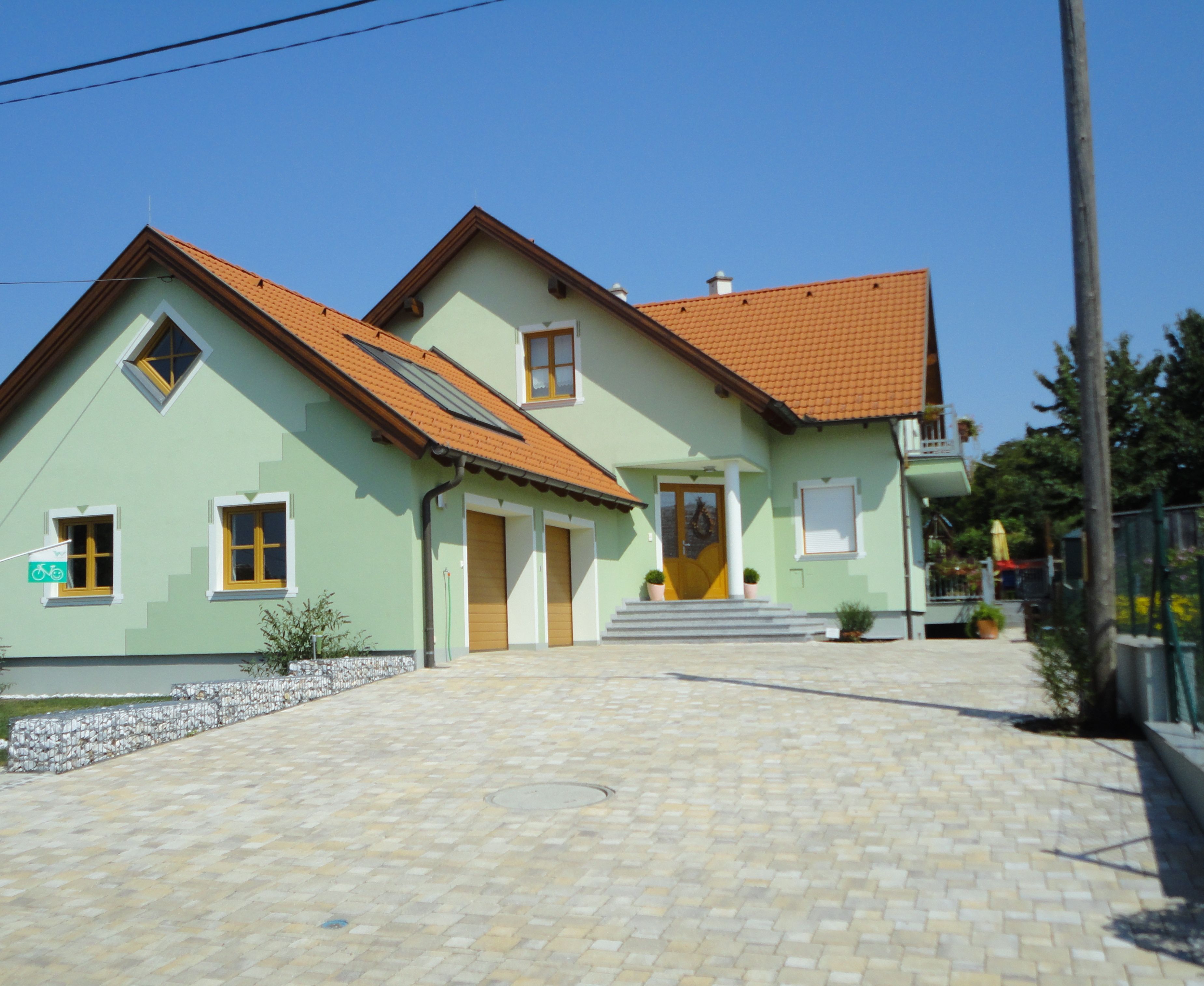 A modern, two-story house with green paint and red roof tiles, surrounded by a paved courtyard.
