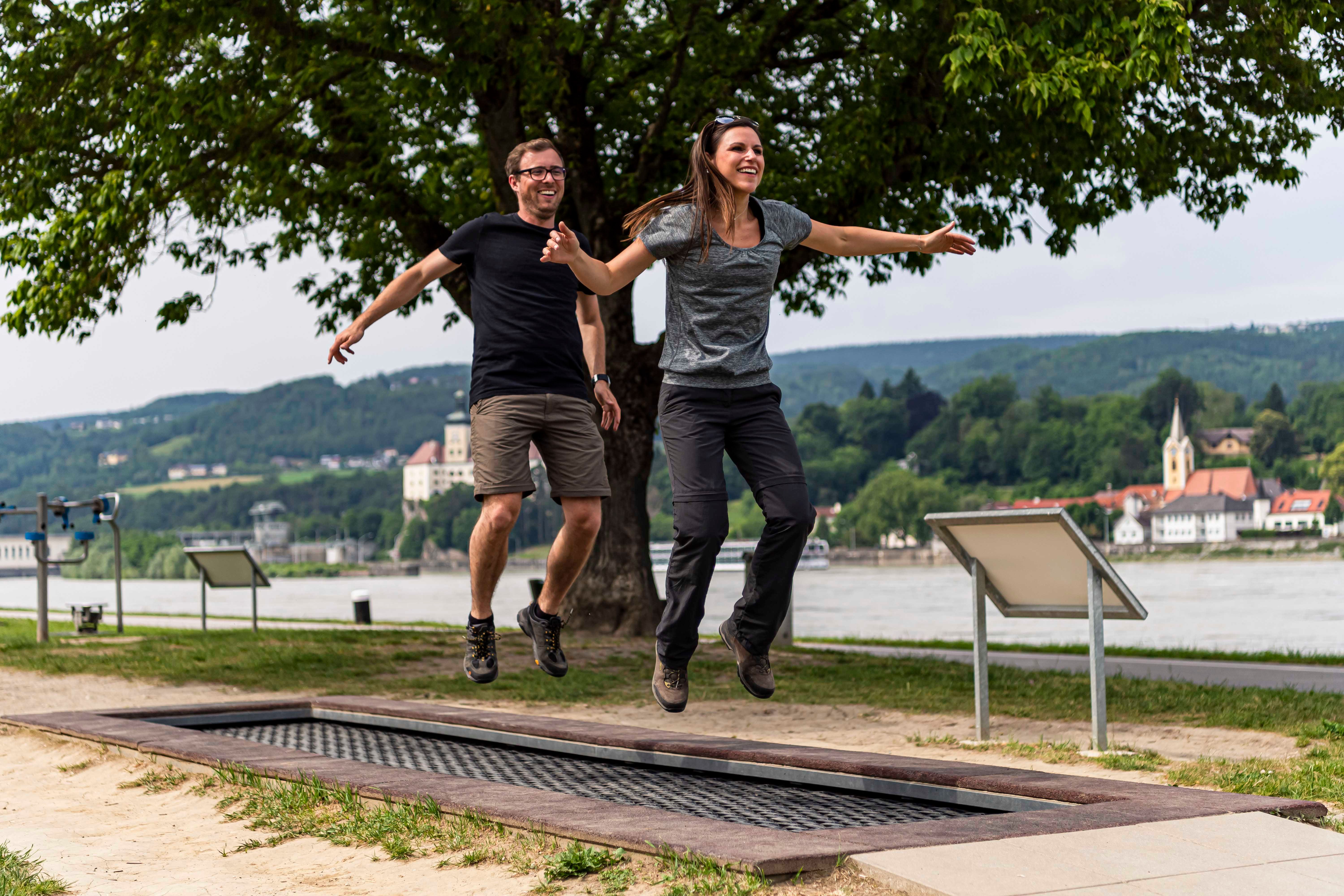 Two people jumping on a trampoline outdoors with a river and buildings in the background.