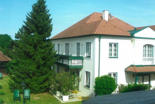 A two-storey white guest house with a red roof and green shutters, surrounded by a garden with trees and garden furniture.