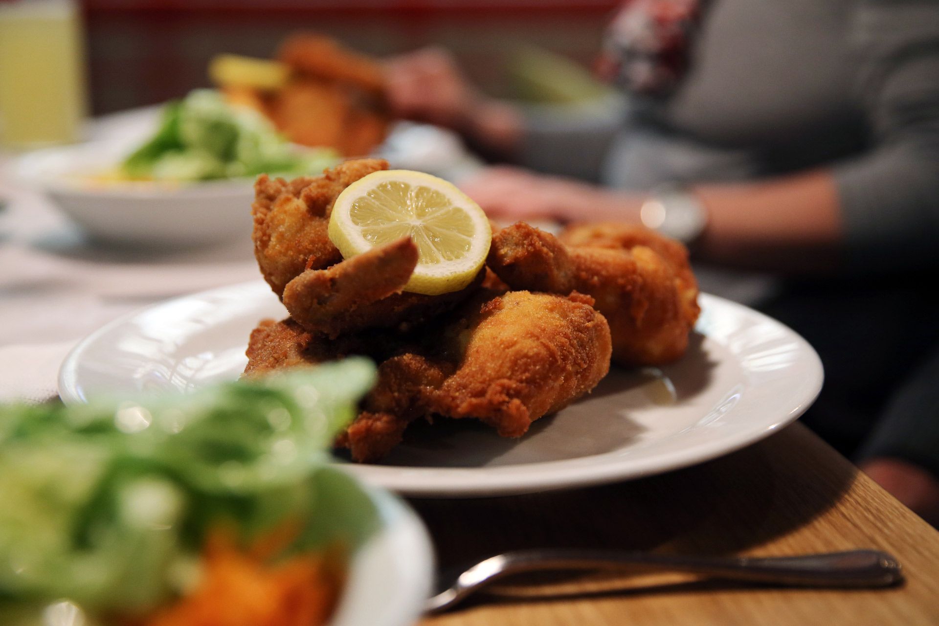 A plate with breaded fried chicken and a slice of lemon, with a person and salad in the background.