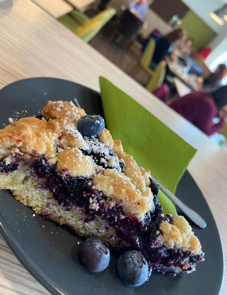 A slice of blueberry cake on a black plate with fresh blueberries and powdered sugar, with a blurred café scene in the background.