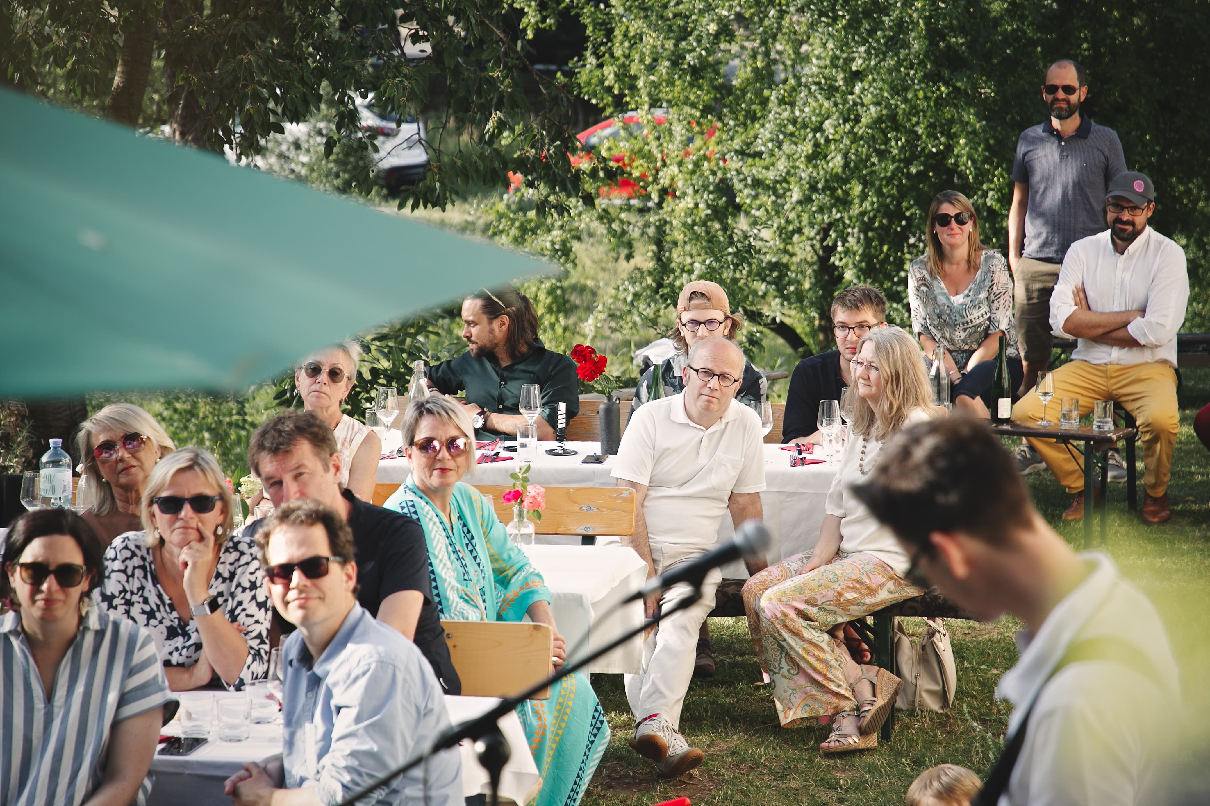 People sitting outside at a concert, surrounded by trees and tables with drinks.