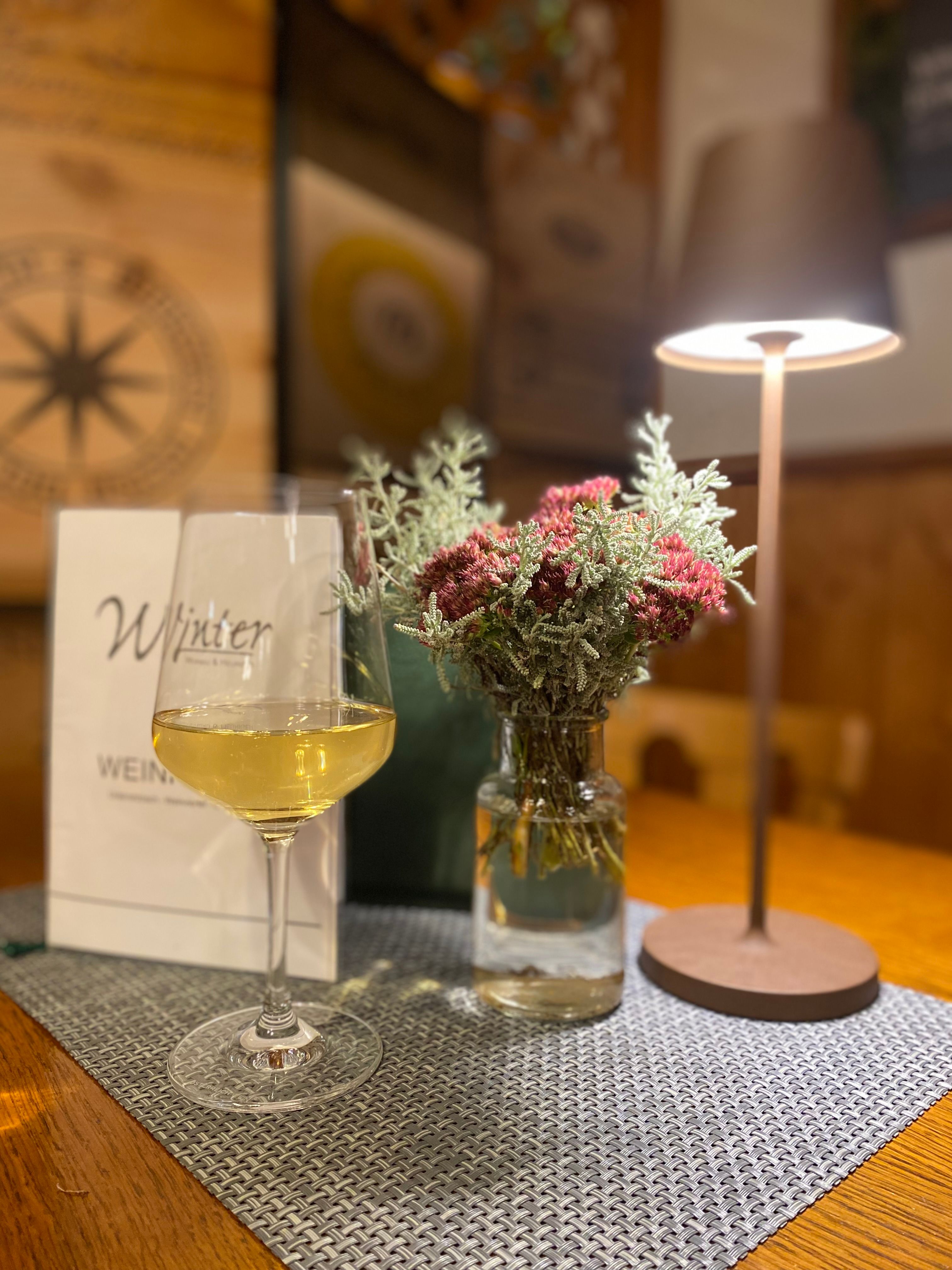 A wine glass with white wine stands on a table next to a vase of flowers and a table lamp.