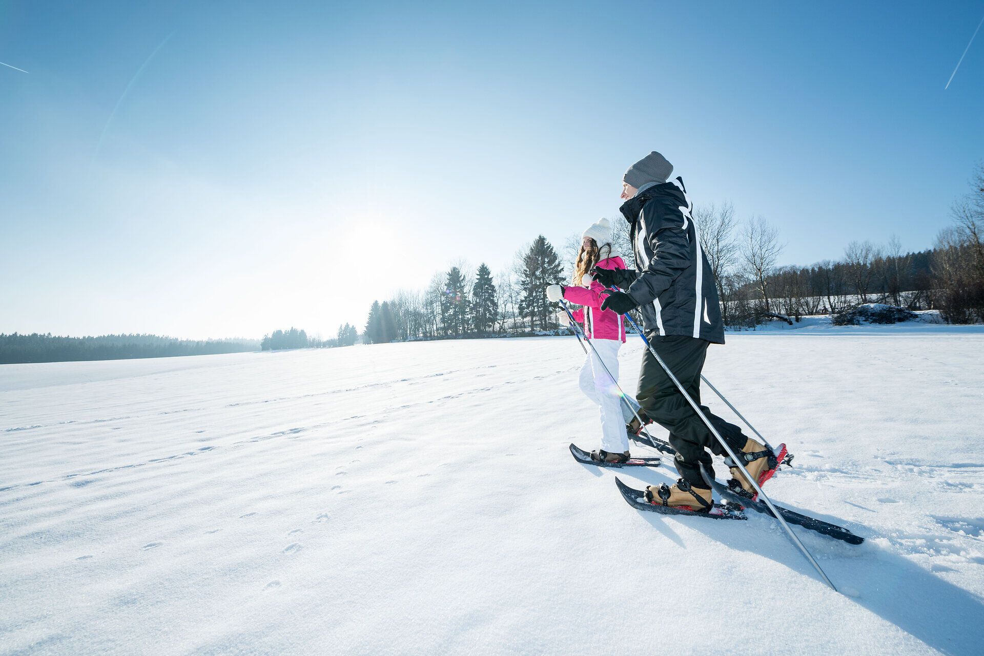 Two people snowshoeing in a snow-covered field under a clear sky.