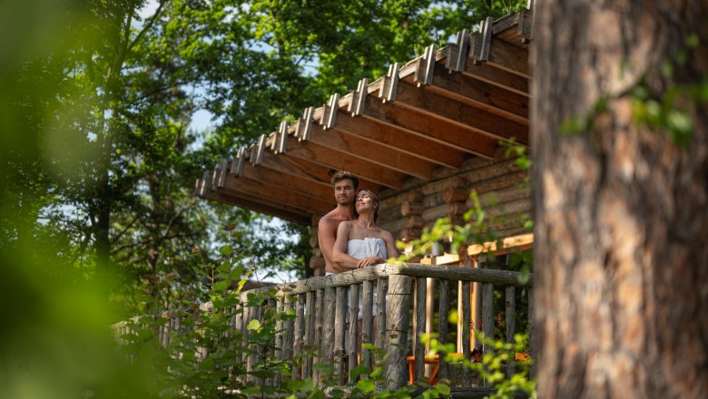 A couple stands on a wooden balcony in a forest, surrounded by trees.
