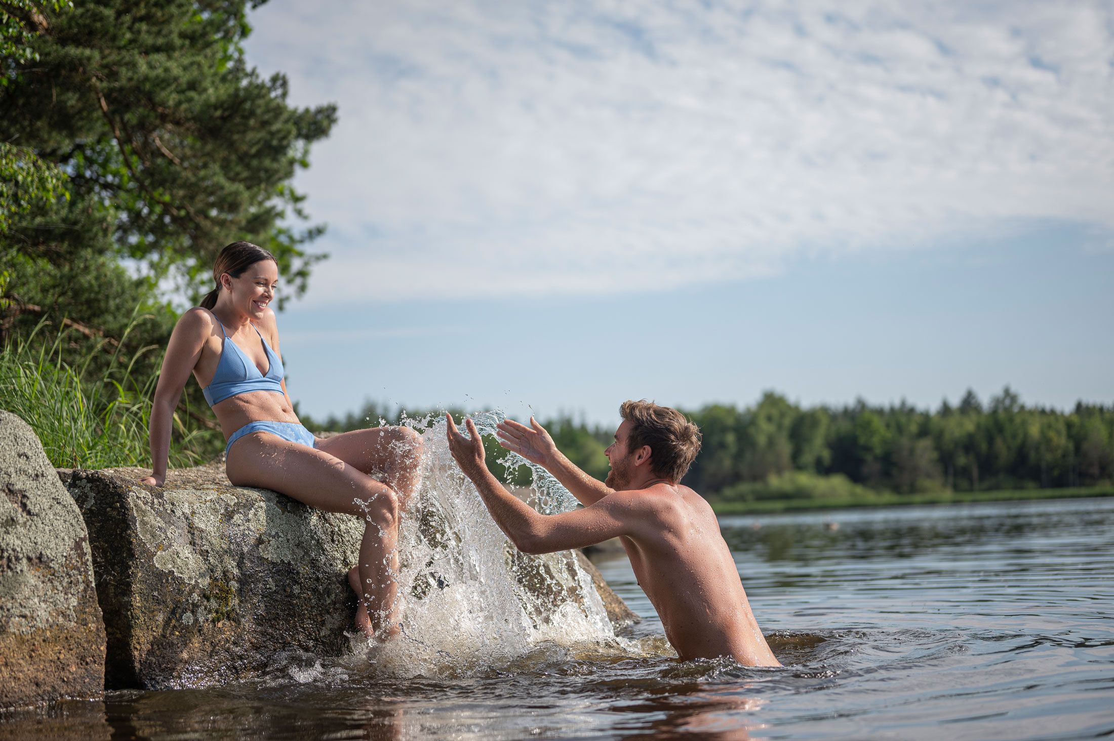 A man in the water splashes a woman sitting on a rock.