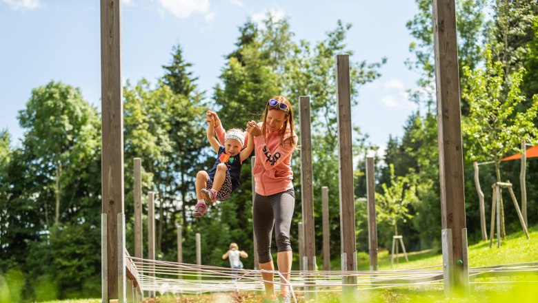 A woman holds a child by the hands in an outdoor play area with wooden posts and ropes.