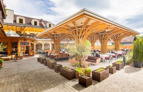 Terrace with wooden roof and tables, surrounded by plants.