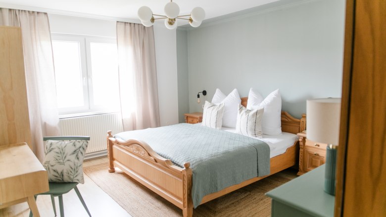 A cozy bedroom with a wooden bed, blue comforter, bedside tables and a desk with chair. Light-colored curtains and a window are visible.