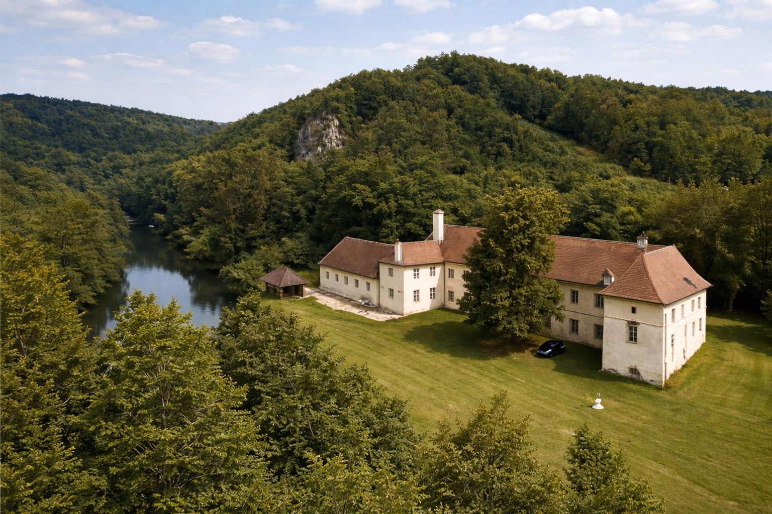 Aerial view of a large, historic building with a red roof, surrounded by forest and a river.