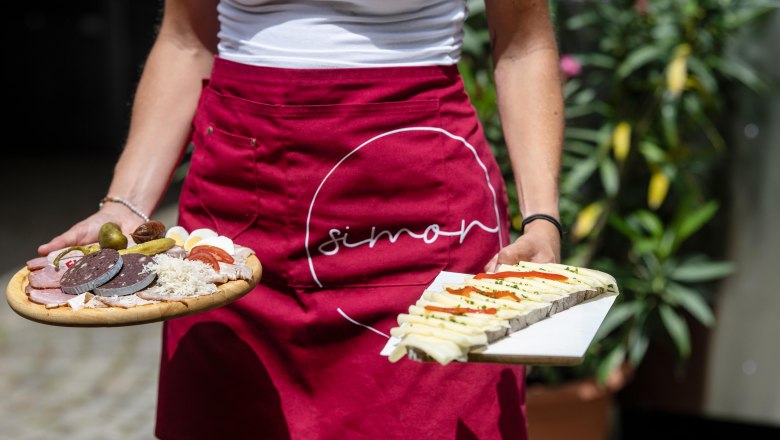 Person in a red apron with the "Simon" logo holds two platters of sausage and cheese.