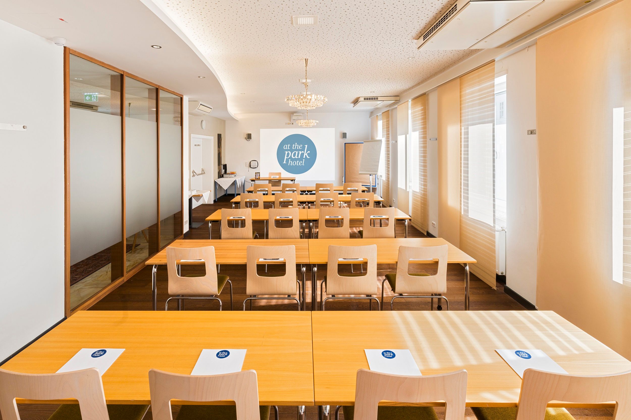 A conference room with tables and chairs, a projector screen and notepads with the "at the park hotel" logo.