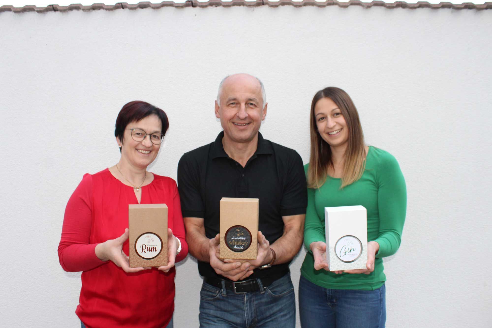 Three people hold gift boxes with rum, whiskey and gin in front of a white wall.