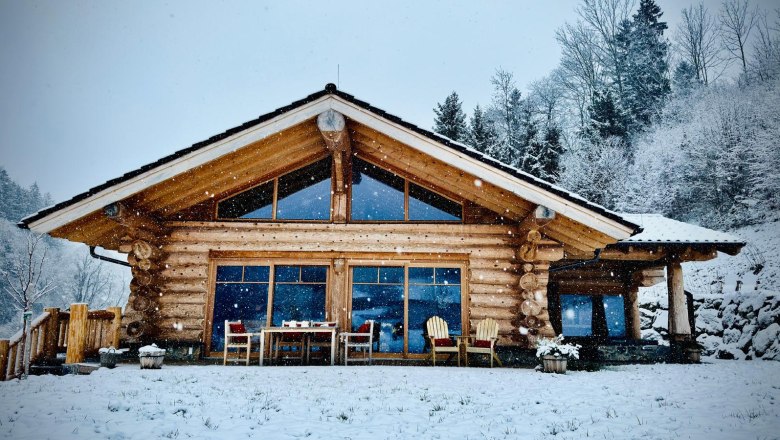 Log cabin in the snow with large windows and terrace, surrounded by snow-covered trees.