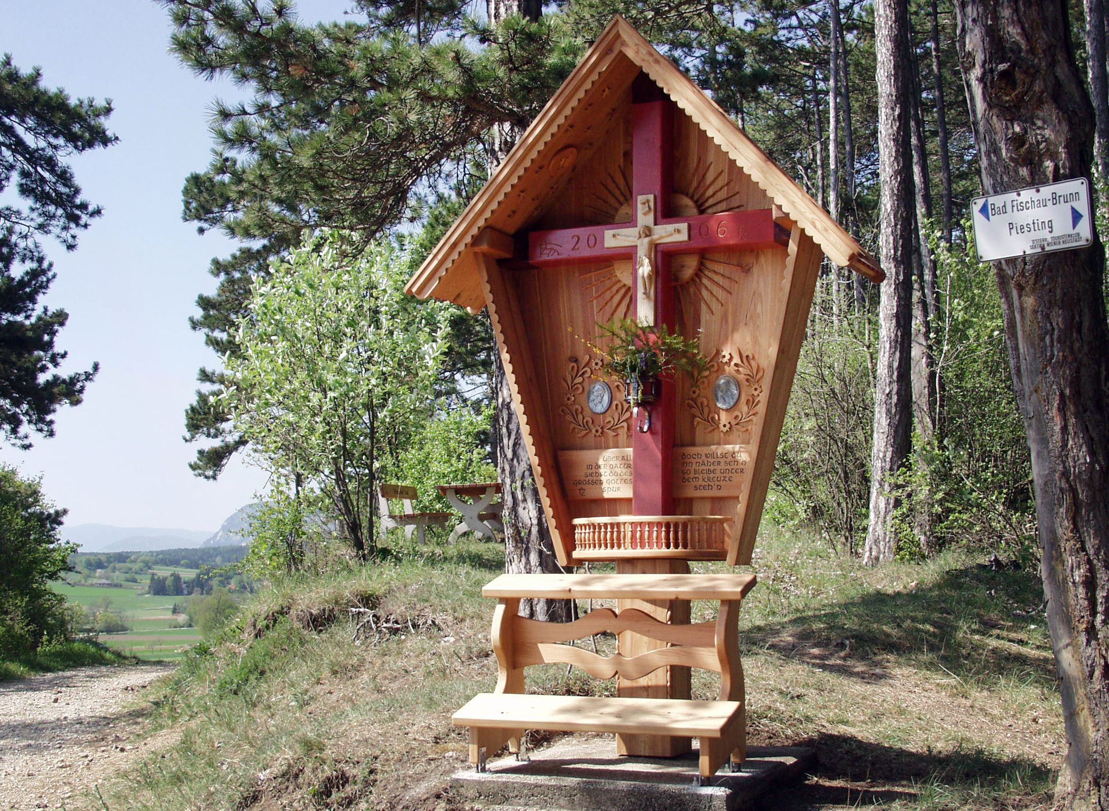Wooden cross with red beam and carved roof in a forest landscape.