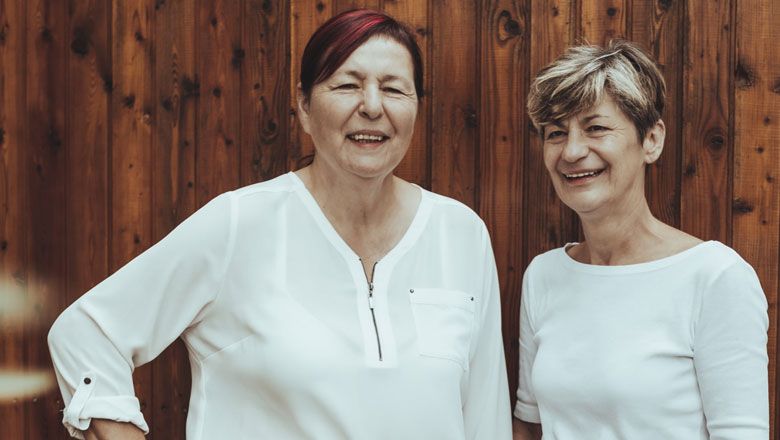 Two women in white blouses stand in front of a wooden wall and smile.