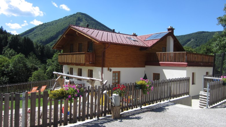 A two-storey house with a wooden balcony and garden, surrounded by mountains and trees.