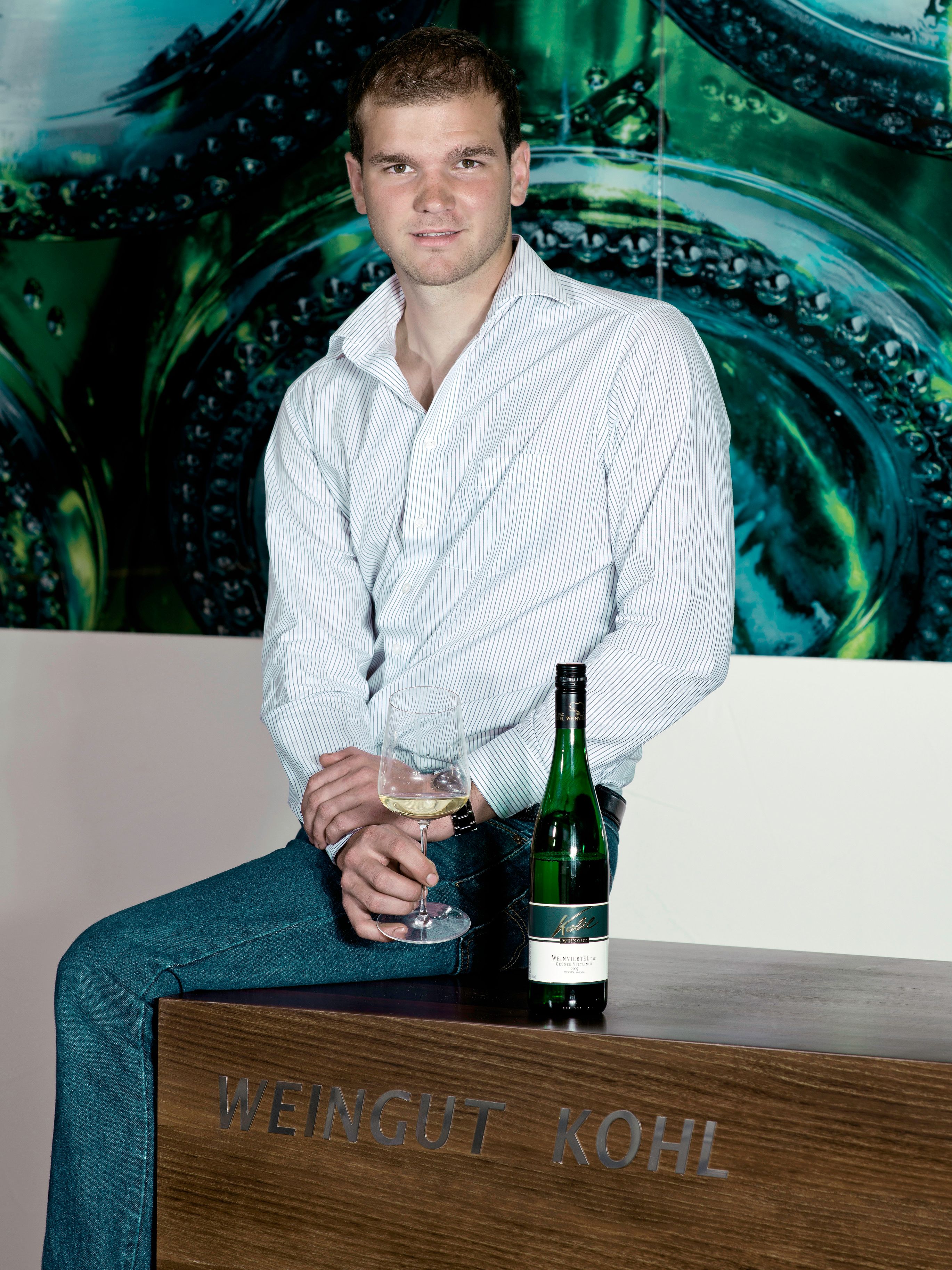 A man is sitting on a table labeled 'Weingut Kohl', holding a wine glass and a wine bottle next to him.