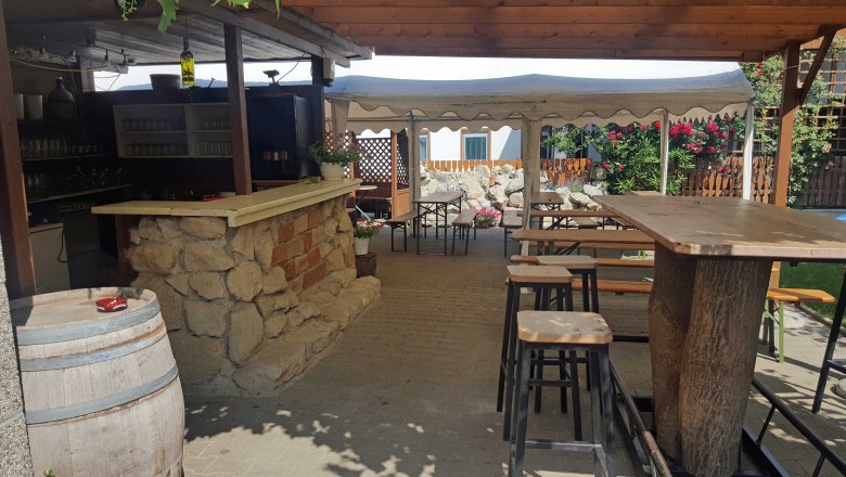 Cozy outdoor area of a Buschenschank (typical tavern) with wooden tables and benches, bar made of stone and wood, surrounded by flowers and plants.