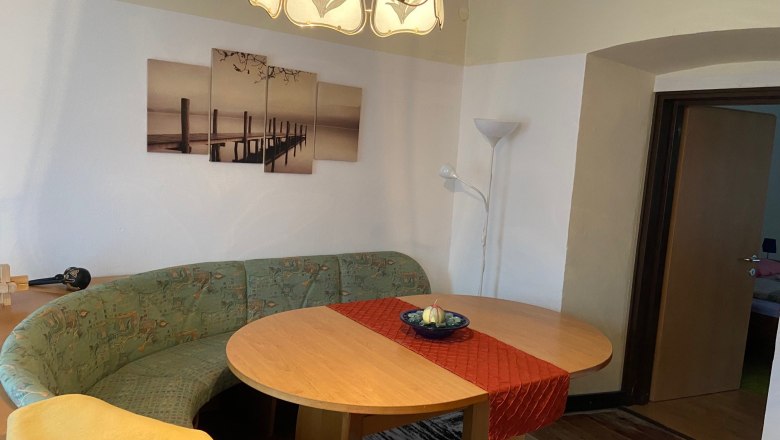 A living room with an oval wooden table, a green corner bench, a red table runner and a picture on the wall. A floor lamp stands in the corner.