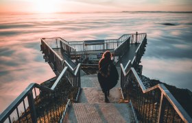 A person walks into the sunset on a skywalk above a sea of fog.