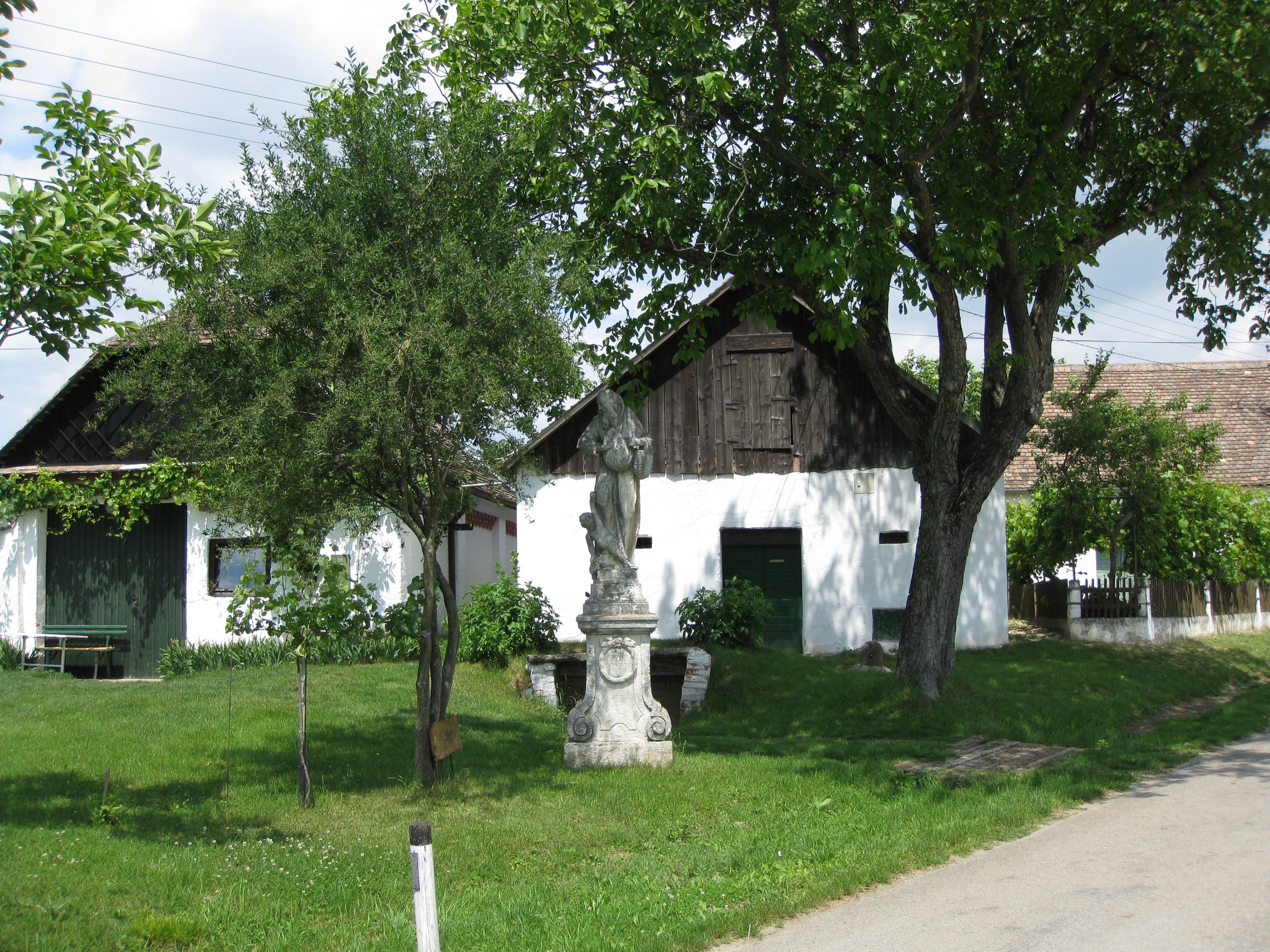 A white building with a wooden gate and statue in the foreground, surrounded by trees and green lawns.