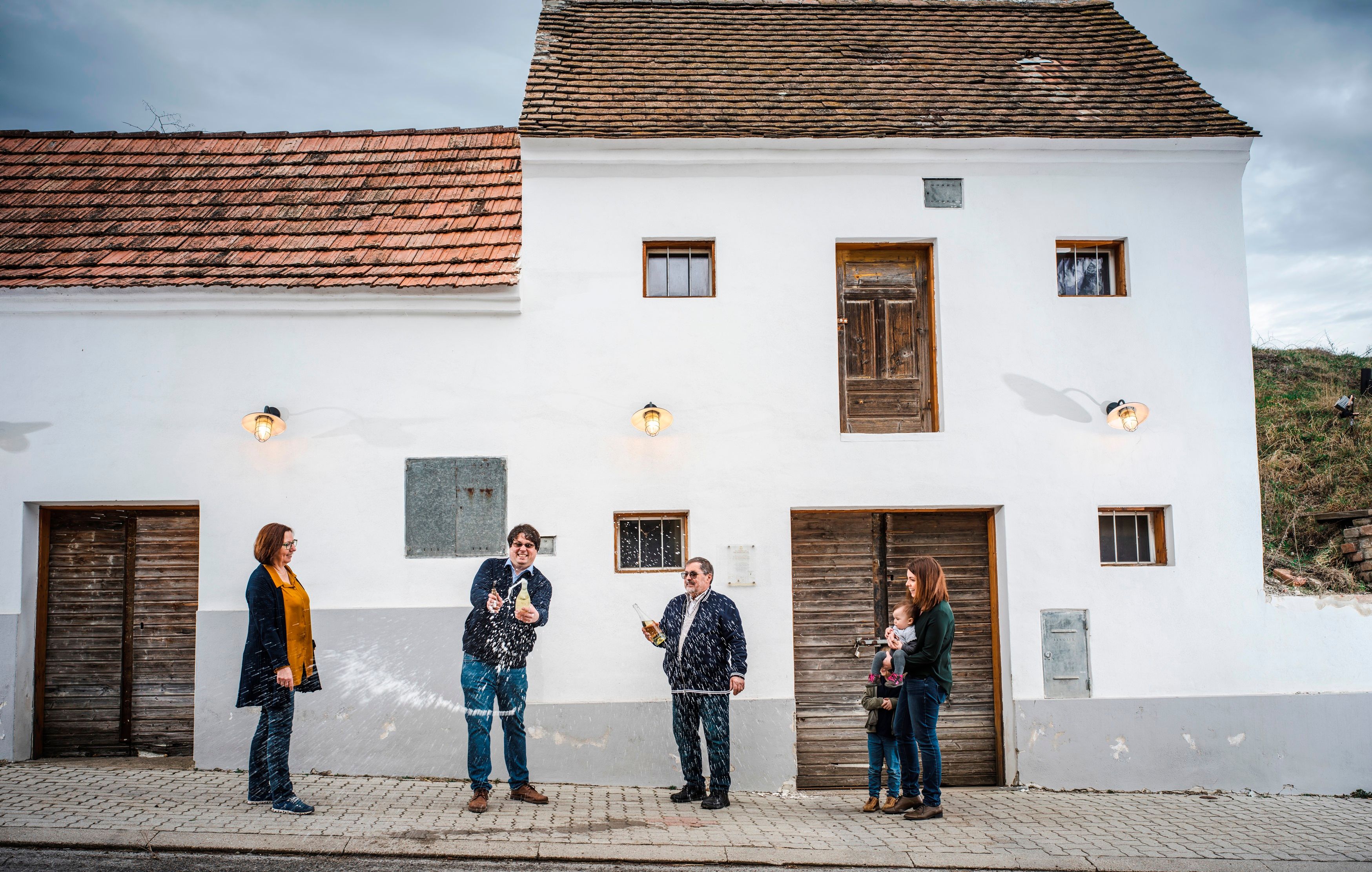 A group of people stand in front of a white building while one person sprays a bottle.