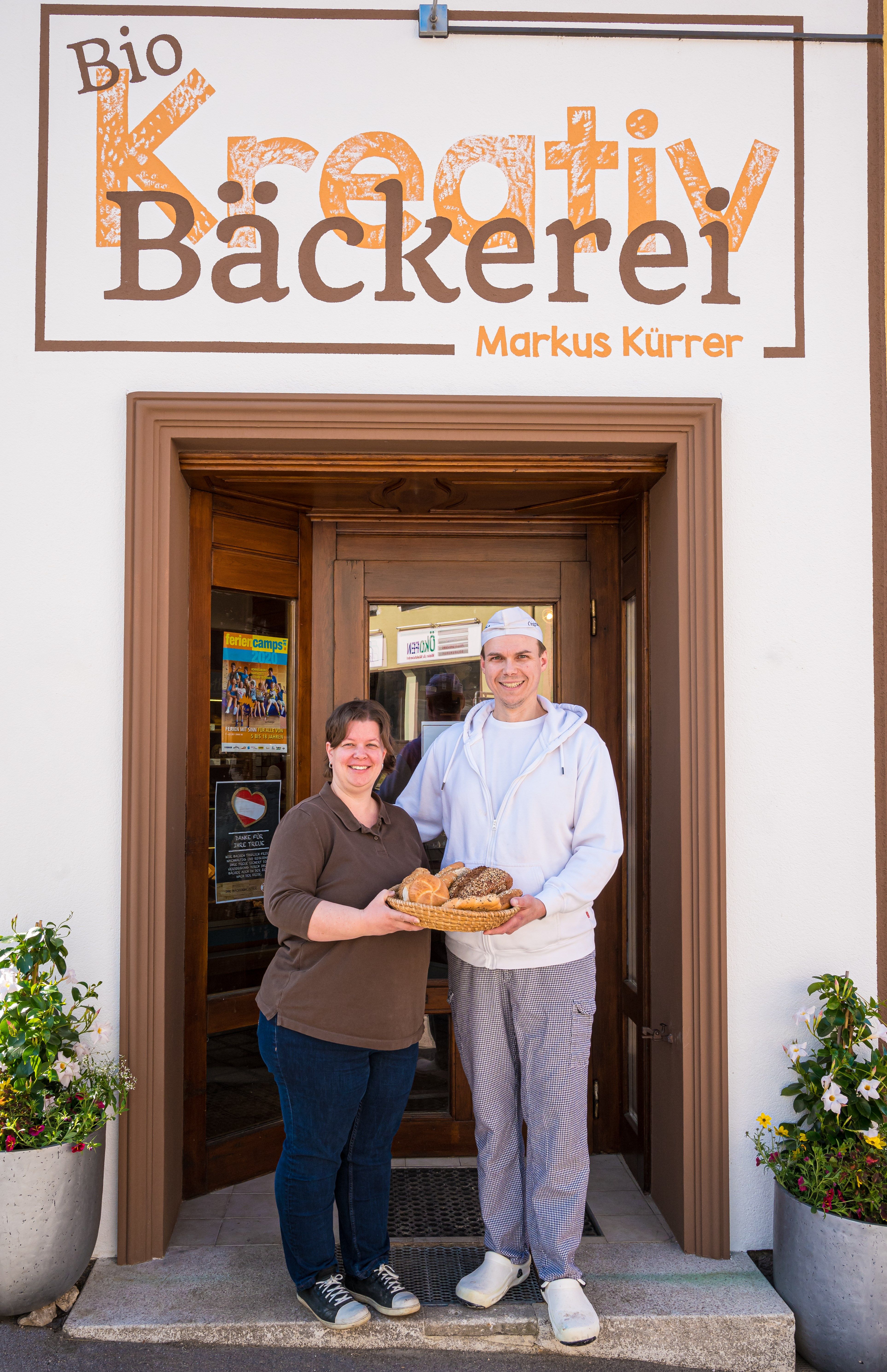 Two people stand in front of Markus Kurrer's Bio Kreativ bakery holding a basket of baked goods.
