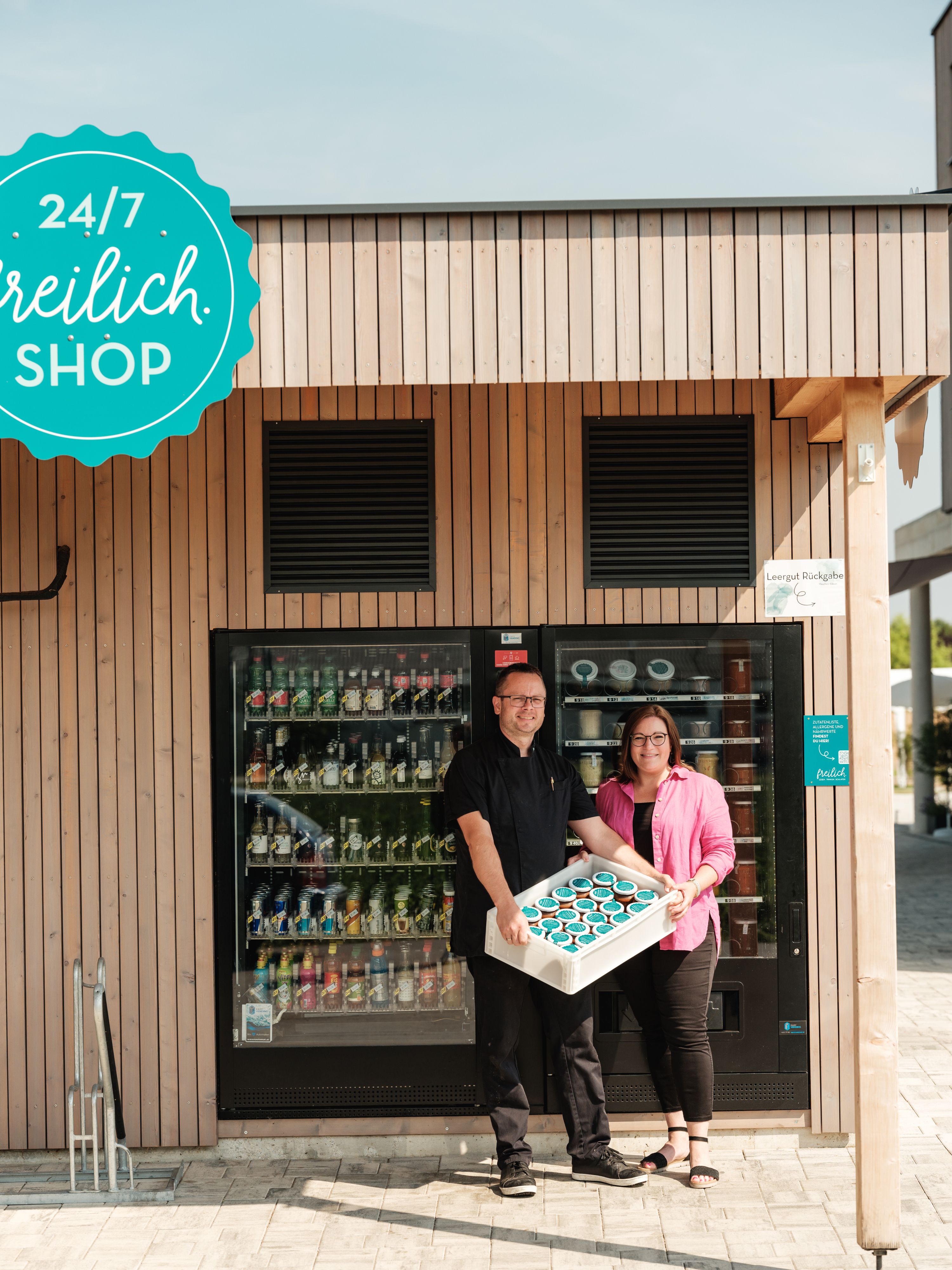 Two people stand in front of a 24/7 open-air store with a tray full of products.