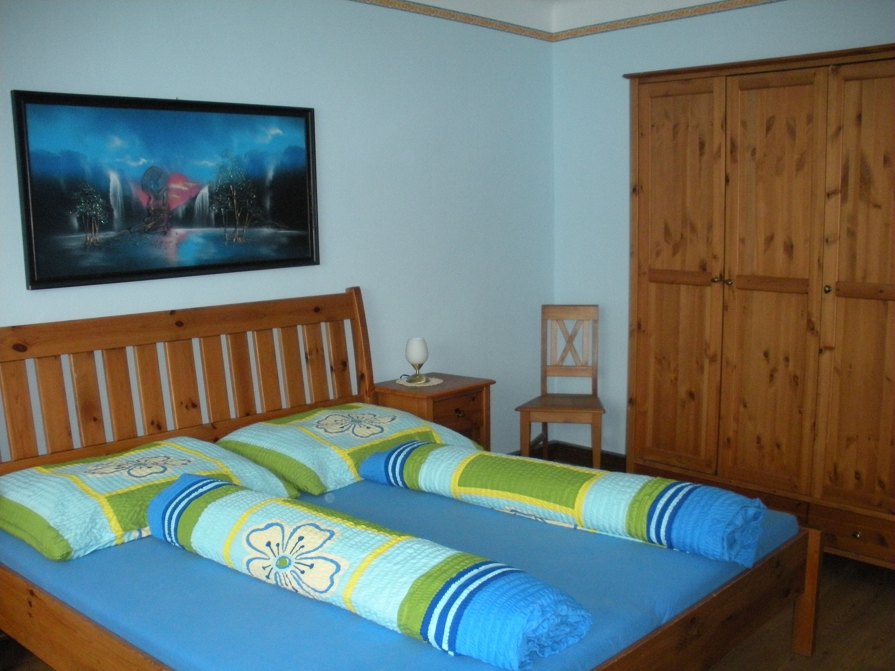 A guest room with a wooden bed, colorful comforters, a wardrobe and a picture on the wall.