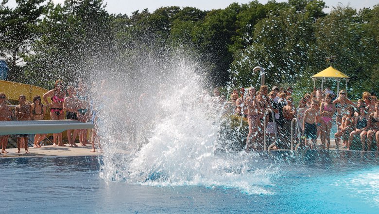 People in an outdoor pool watch a large splash of water in the pool.
