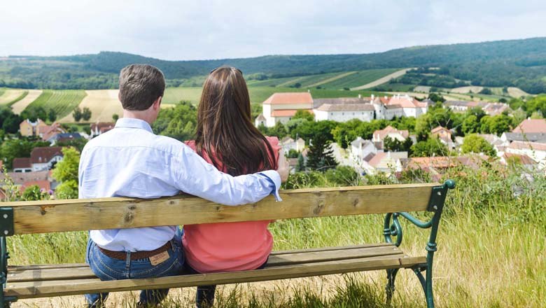 A couple sits on a bench and looks out over a village with a castle in the background.