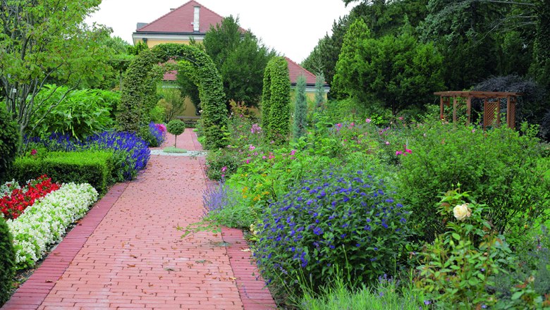 A well-kept garden with a red brick path, surrounded by colorful flowers and green bushes, leads to a house with a red roof.