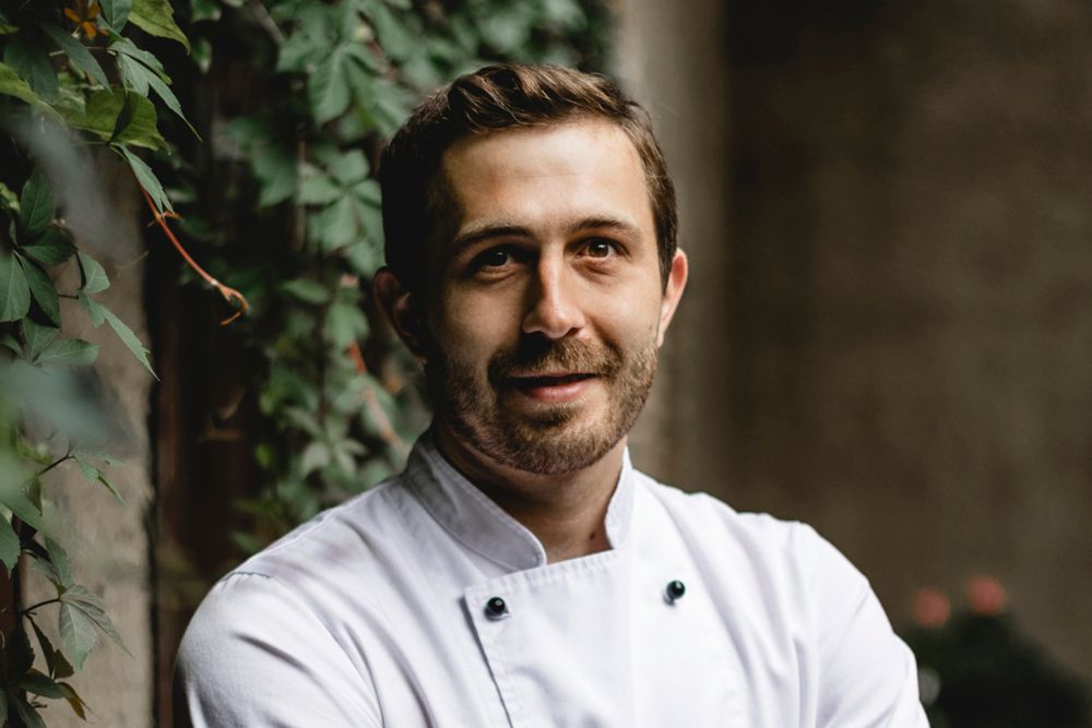 A man in a white chef's jacket stands in front of a wall overgrown with ivy.
