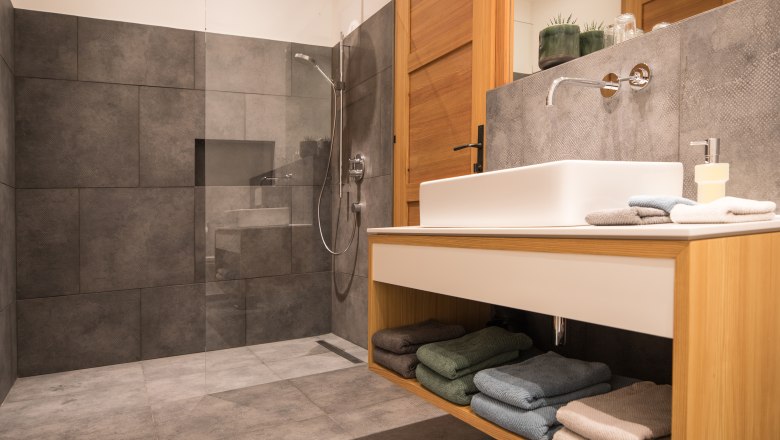 Modern bathroom with gray tiles, glass shower and wooden washbasin.