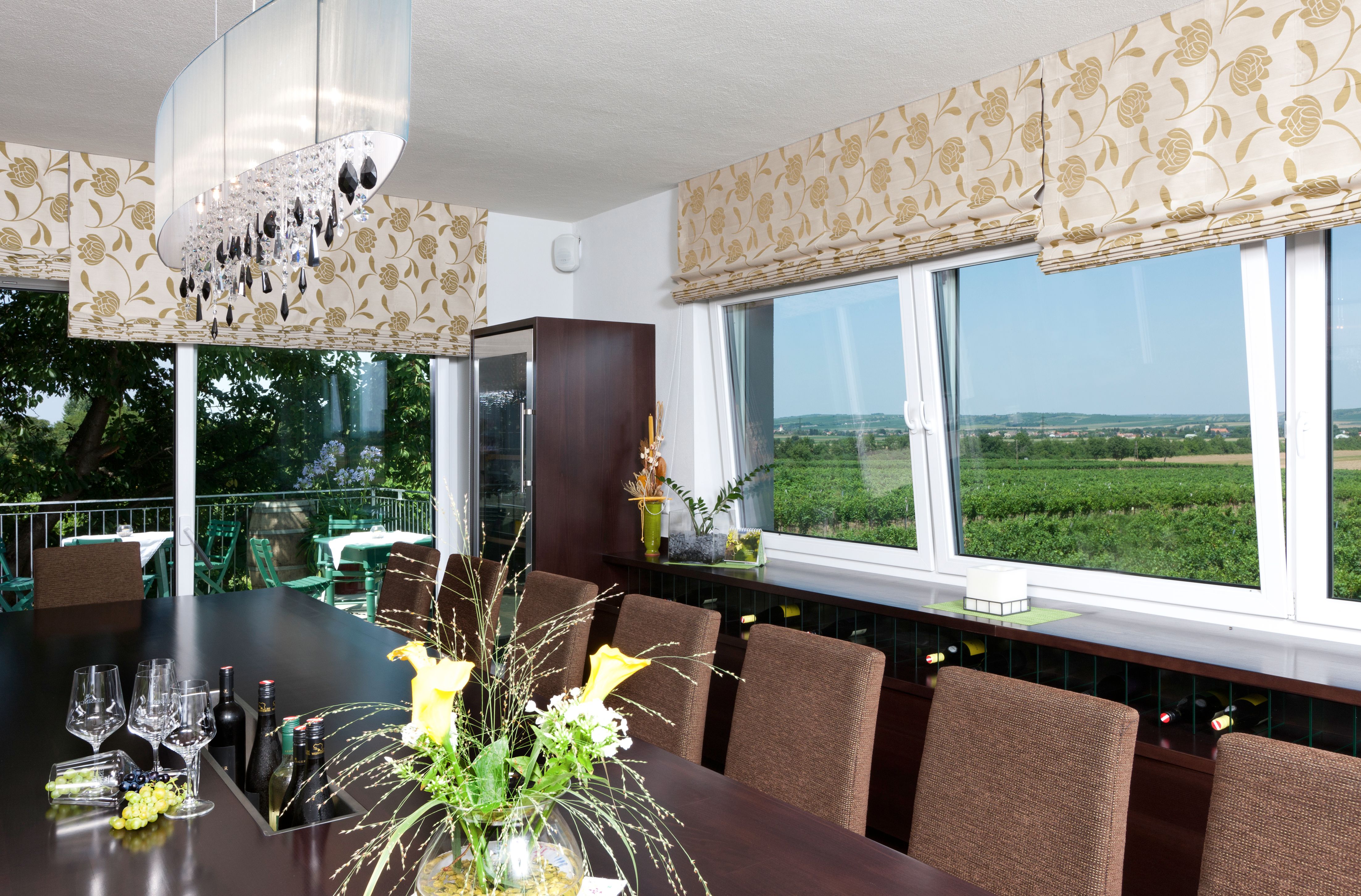 A tasting room with a large table, wine glasses and bottles. Large windows offer views of vineyards. Elegant decoration and lighting.
