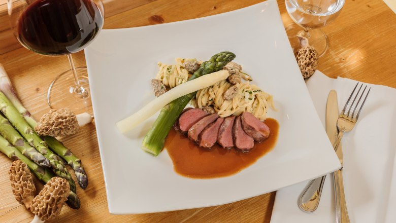 Gourmet dish with meat, asparagus, pasta and morels on a white plate, with a glass of red wine next to it.