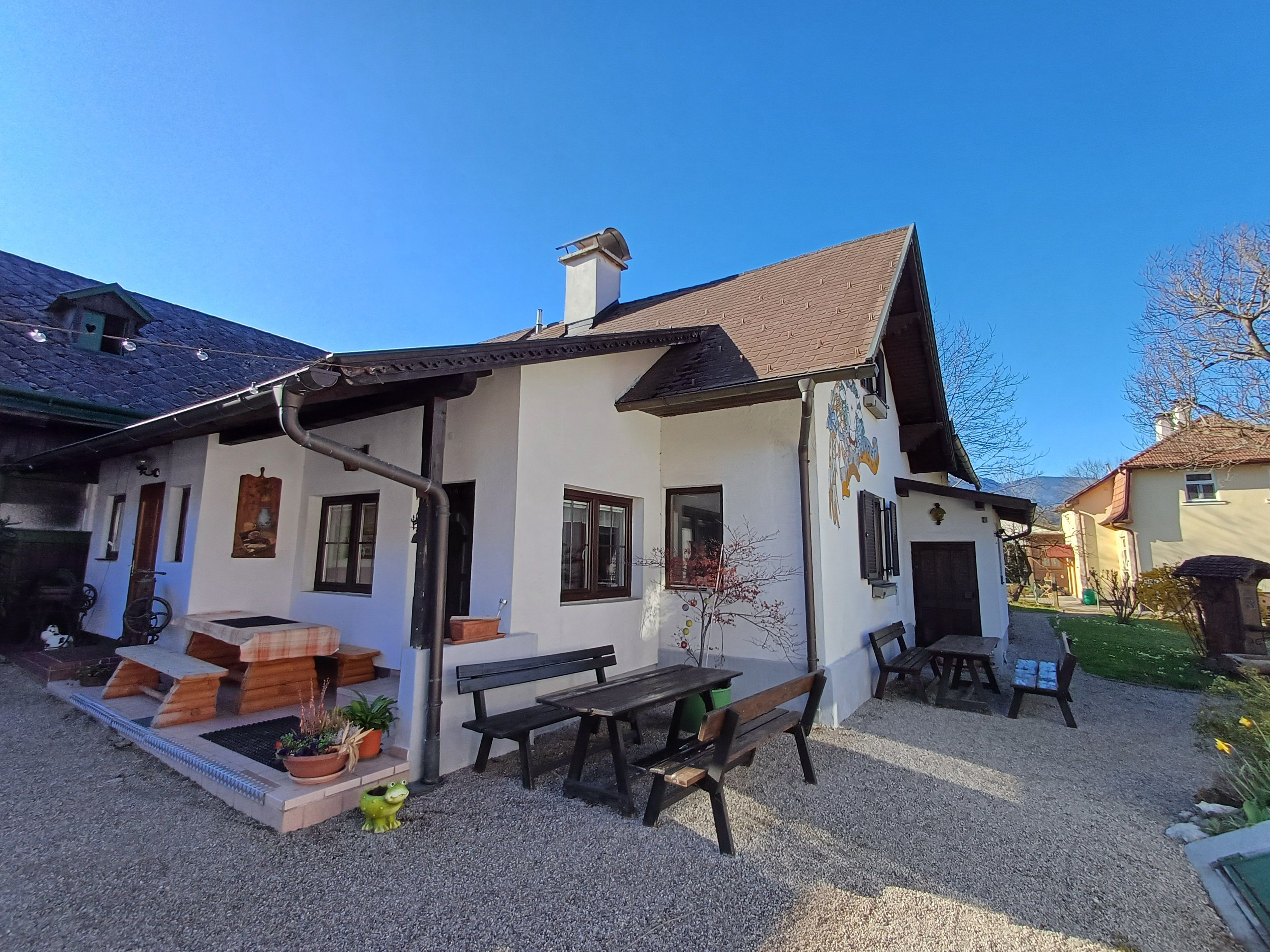 A traditional house with wooden benches and tables in the garden, surrounded by flowering plants and trees under a clear blue sky.