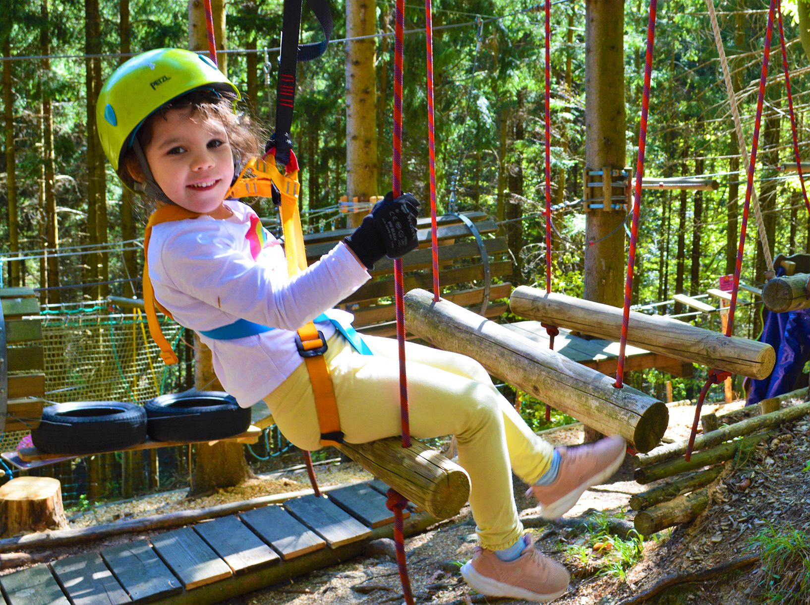 A child wearing a helmet and safety harness sits on a wooden course in the forest.