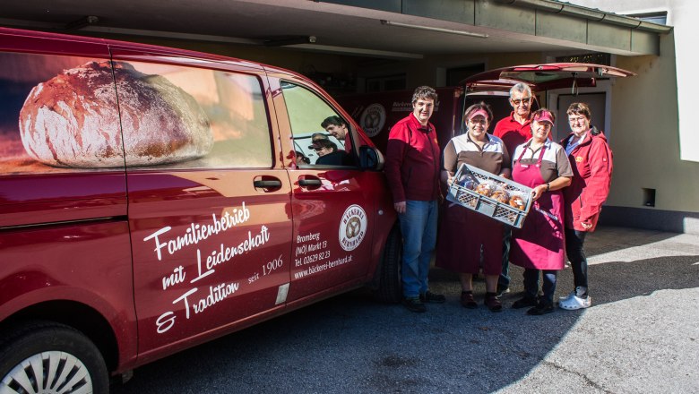 Five people hold a gray box of pastries next to a red van.